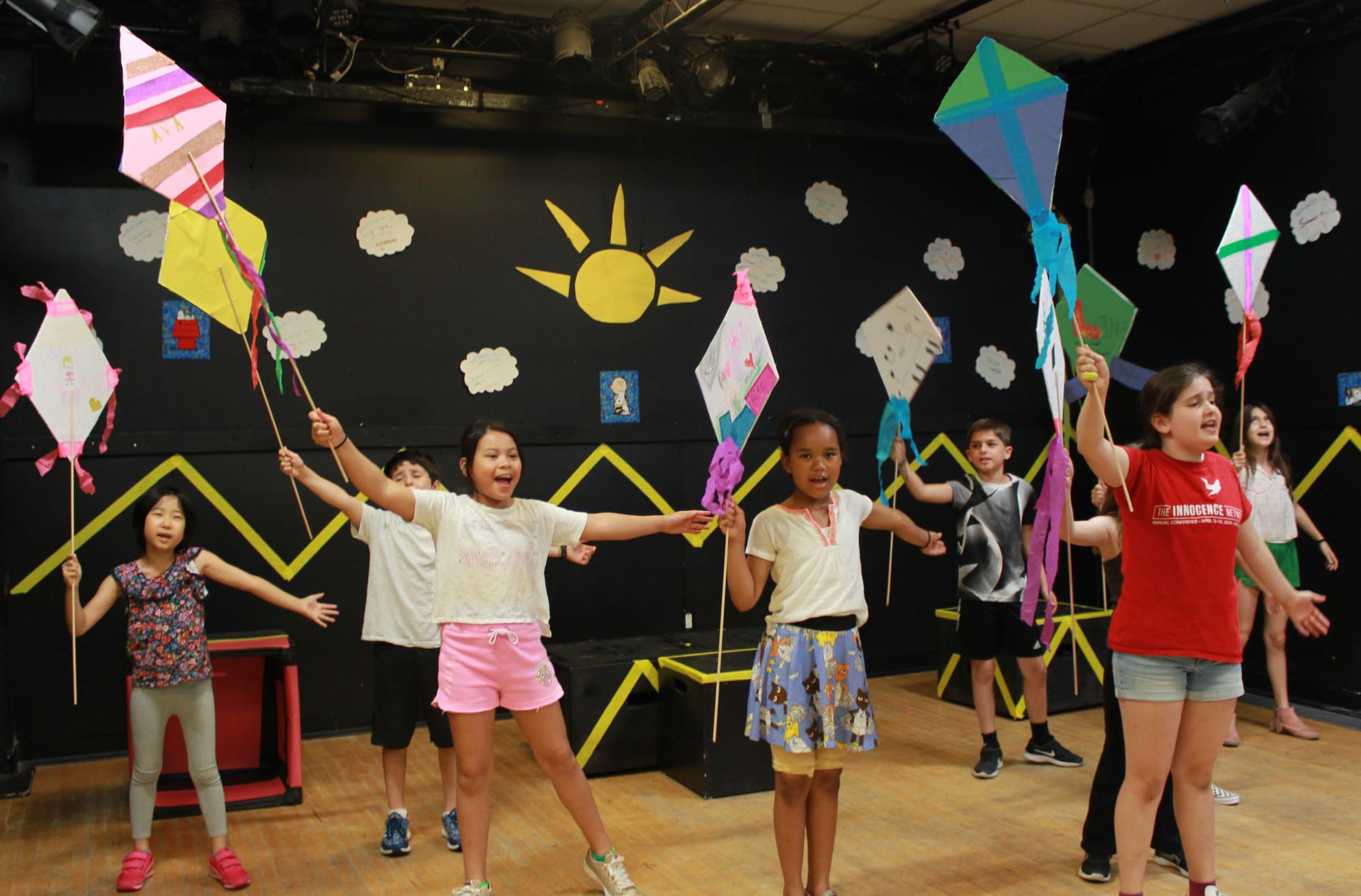 Children perform "We Are in a Play!" with colorful kites on stage.