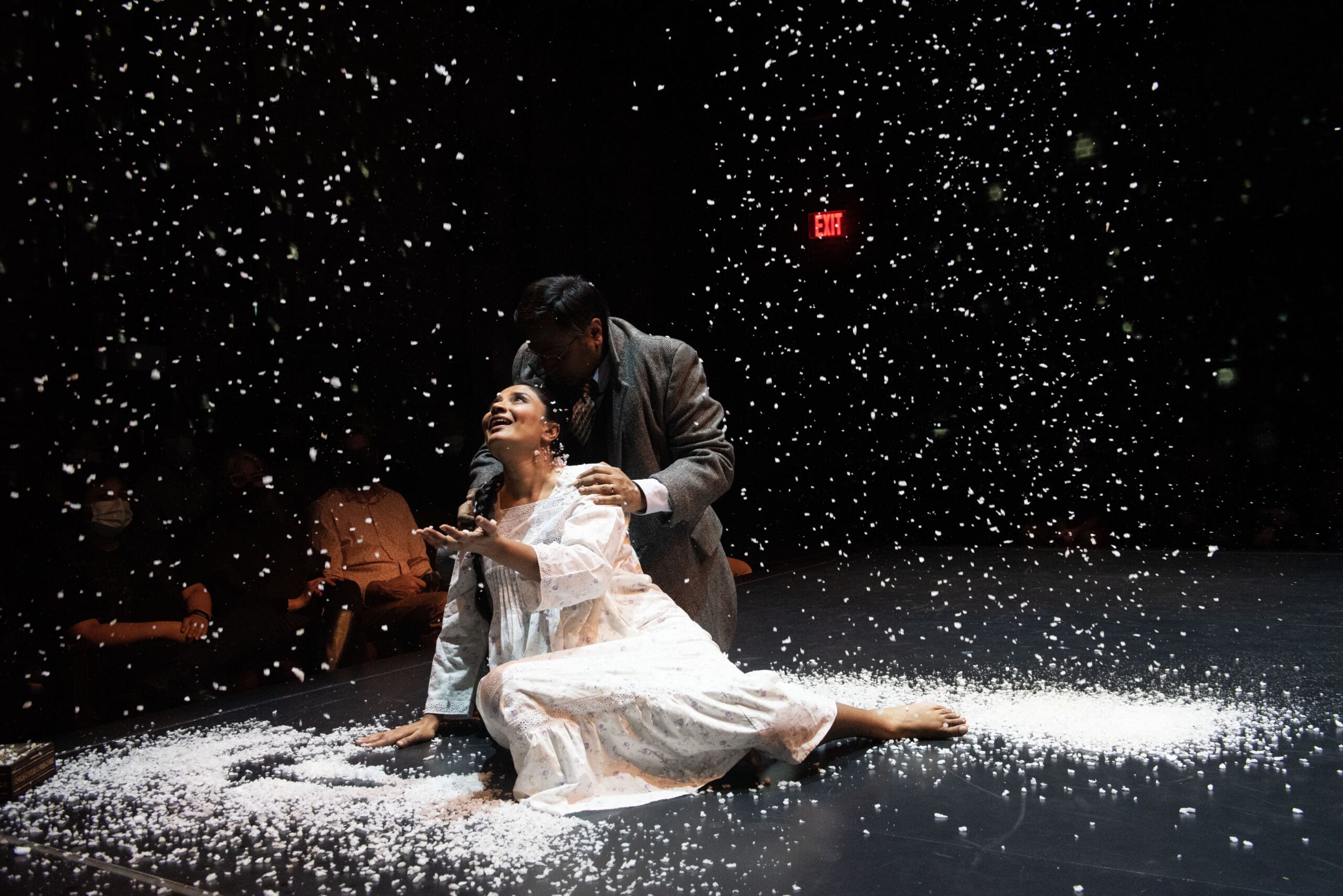 Stage scene with woman in white dress and man in suit under falling snow.