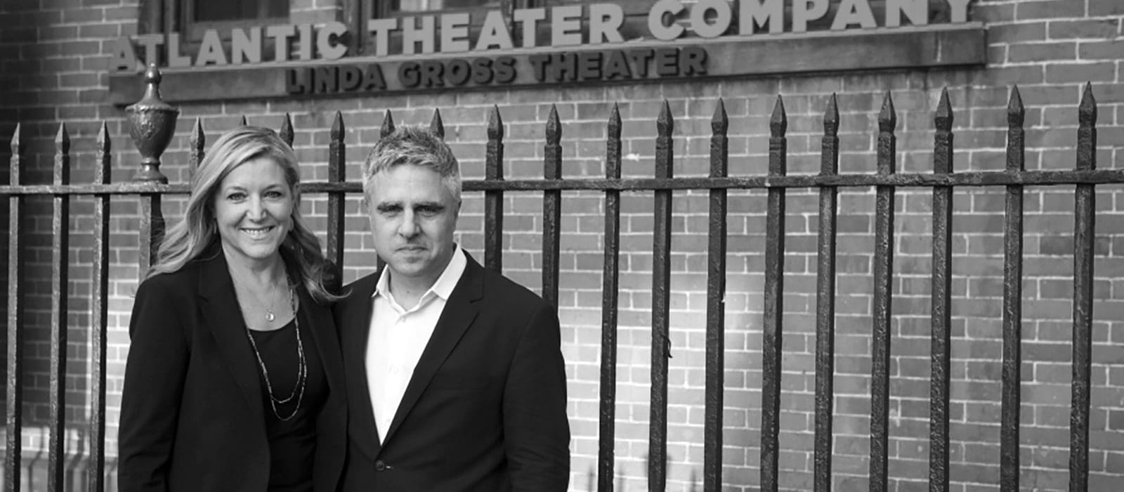 Black and white photo of two people standing in front of the The Linda Gross Theater with the words "Atlantic Theater Company" written on the wall