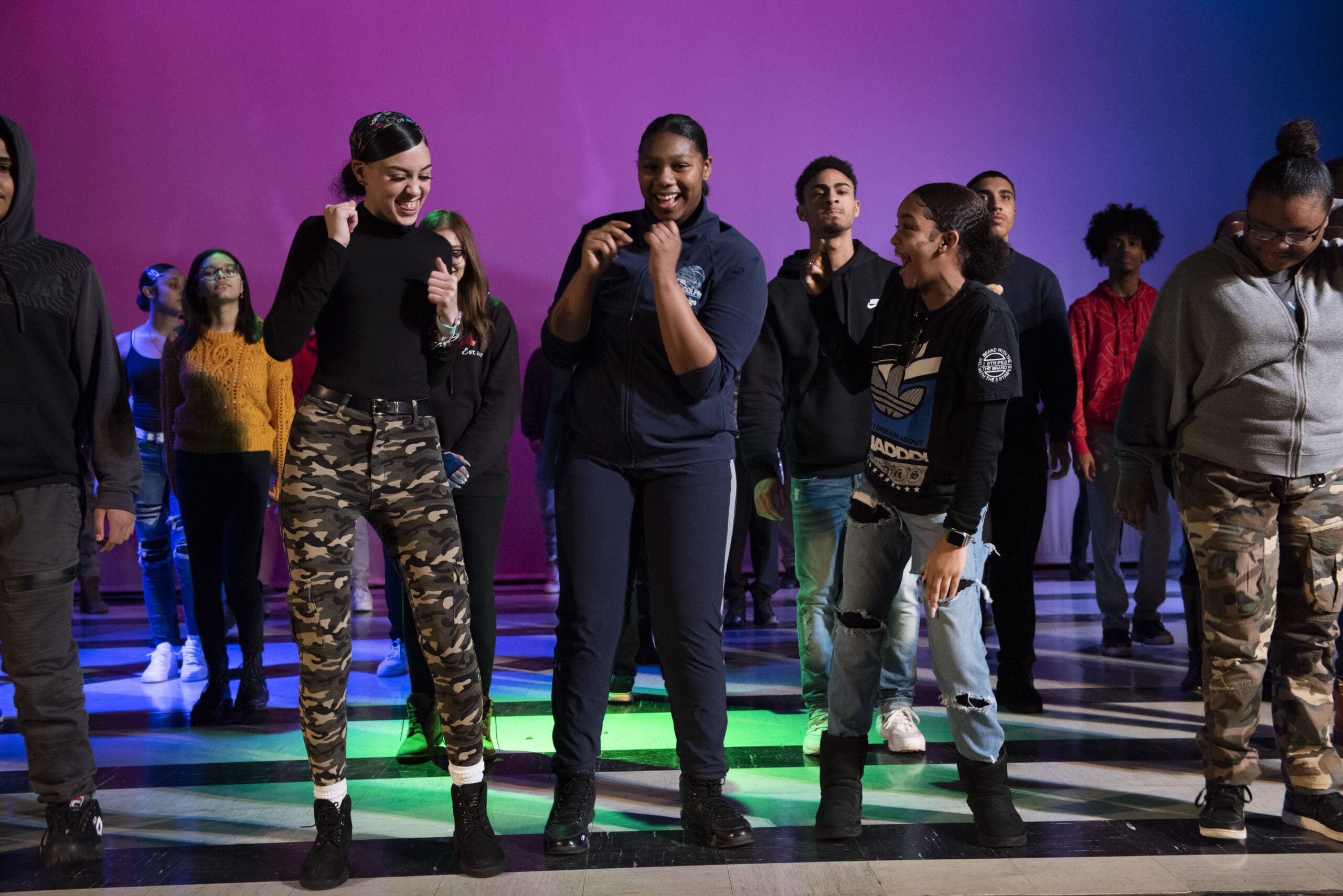 Teenagers performing on stage with a colorful background. Text on a shirt reads "Adidas".