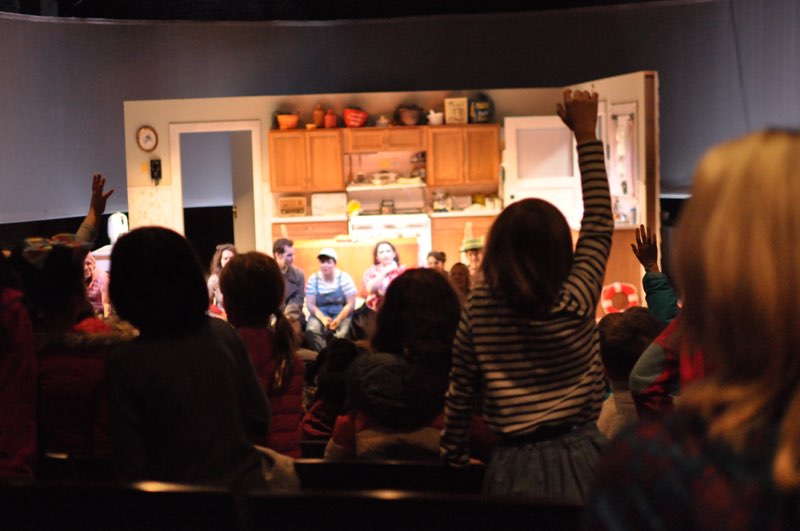 Children raising hands at a Relaxed Performance, focused on a kitchen set.