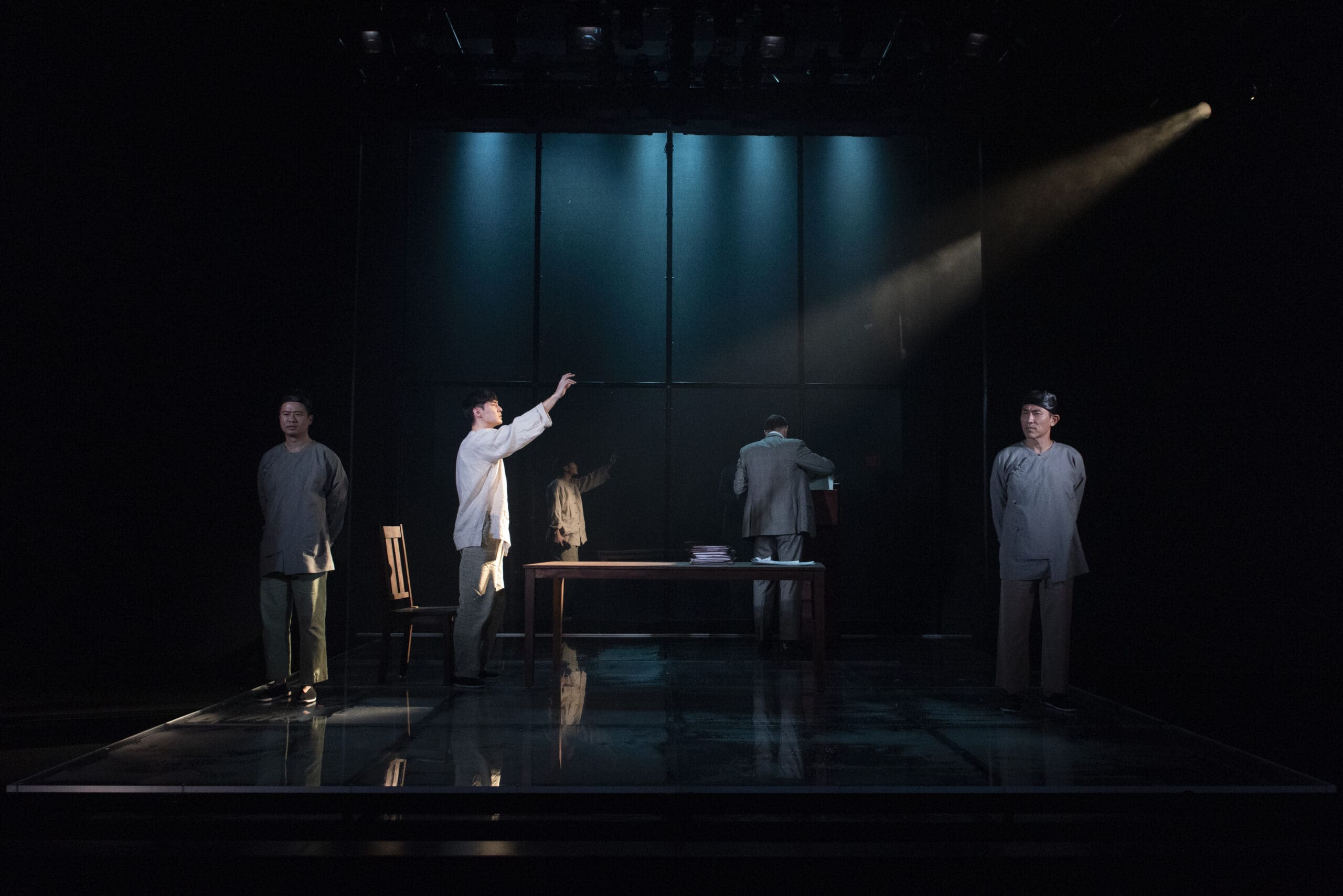 Stage performance with actors around a table under spotlight.