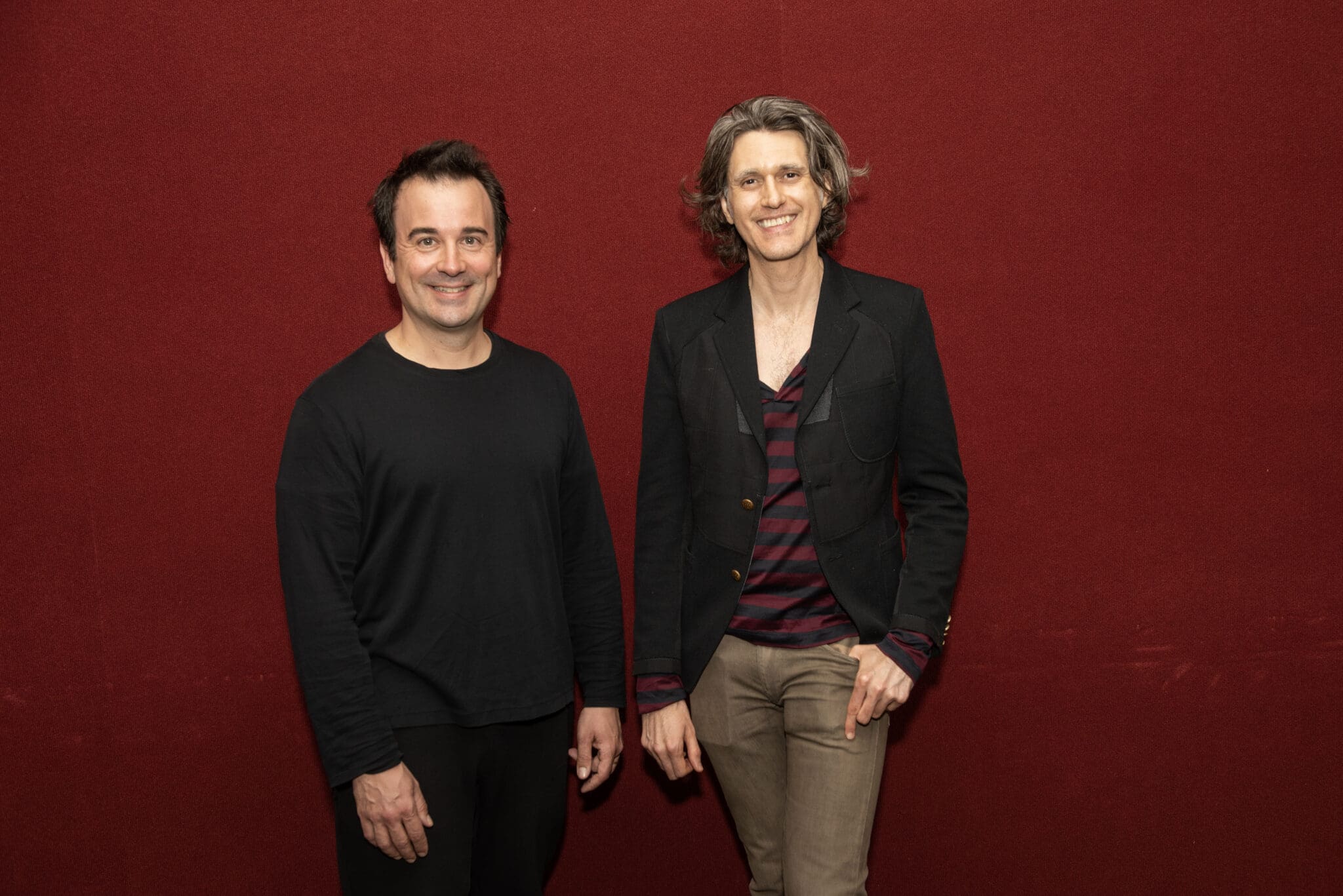 Two men stand against a red background. One wears a black shirt, the other a blazer and striped shirt.