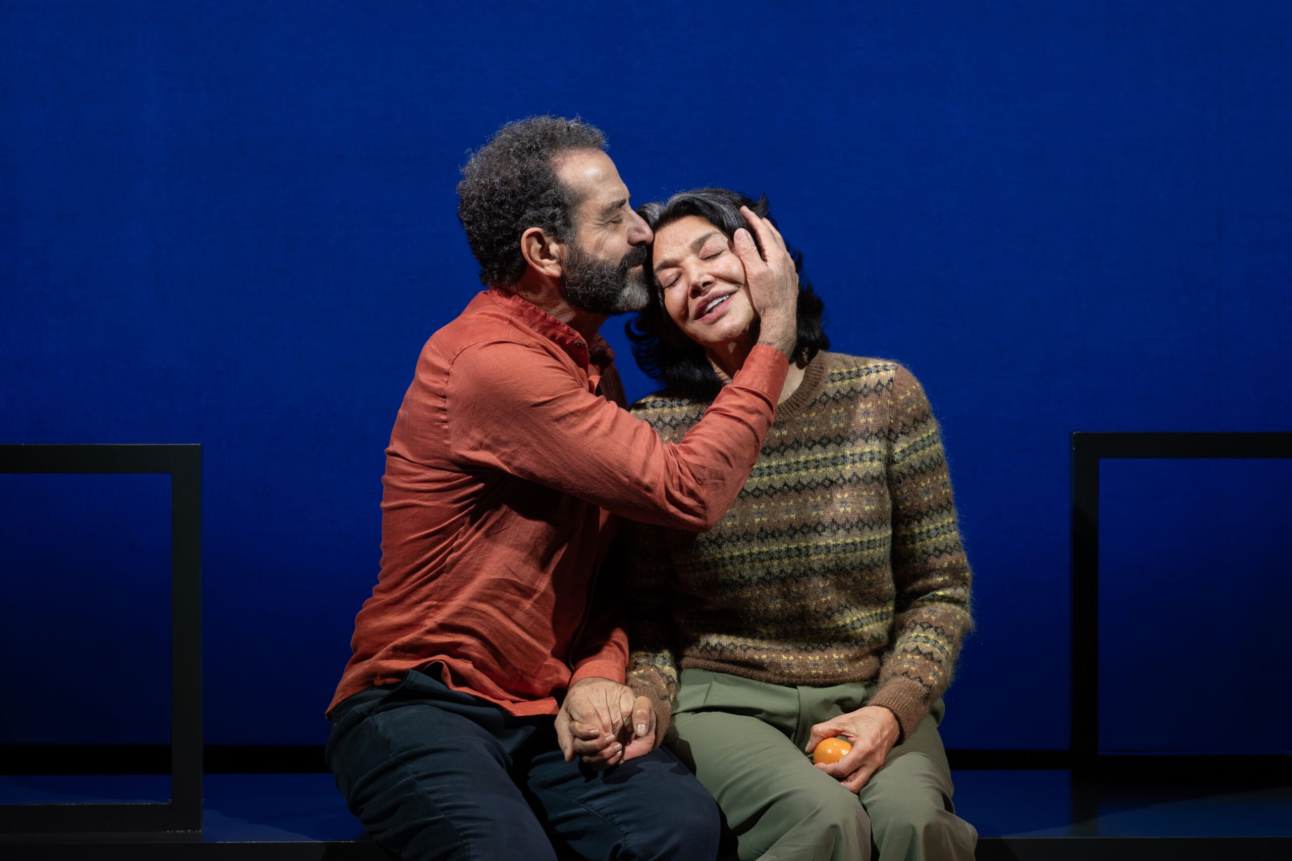 Man kisses woman's head, holding hands, with an orange. Intimate moment on a blue backdrop.