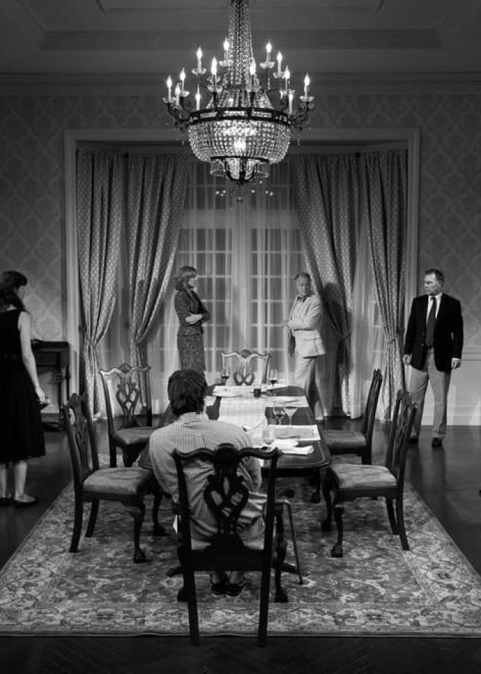 Black and white stage photo of a play, featuring actors around a dining table. The set evokes a sense of drama.