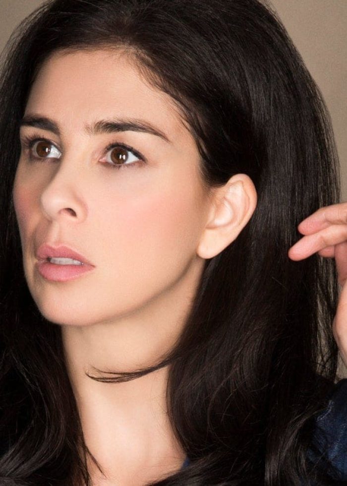 Sarah Silverman looking thoughtful, dark hair, hand near ear.
