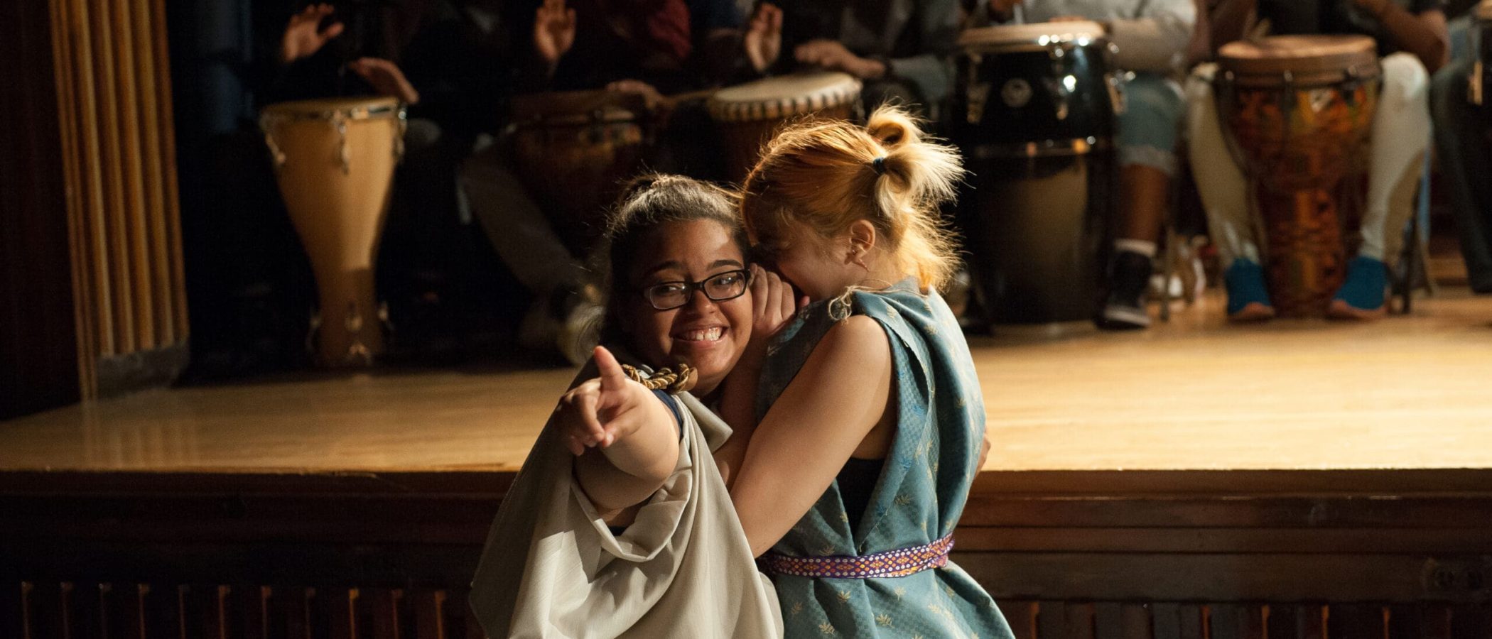 2106SSJRsRehearsal.606 Two young women on stage, smiling and pointing, with a drum circle in the background.