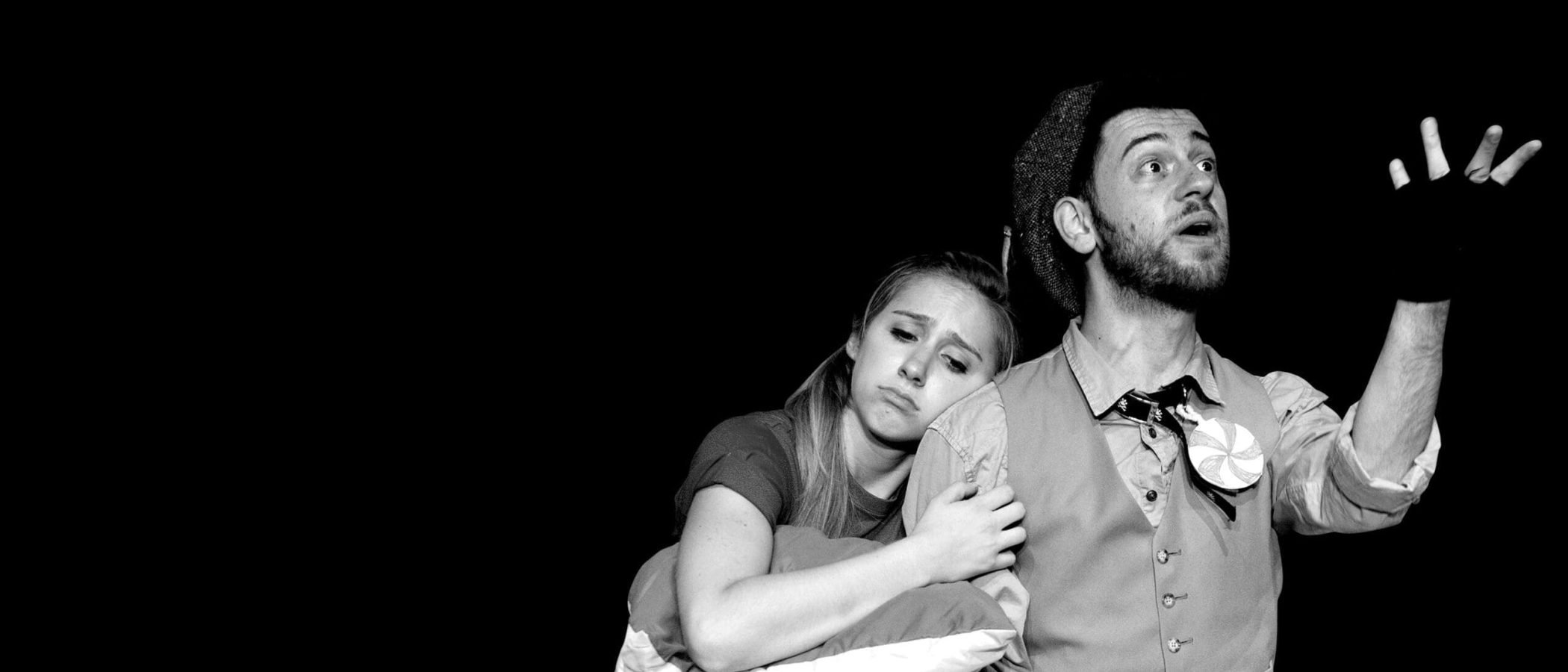 Black and white photo of a man and woman in theatrical costumes. The woman is hugging a pillow while the man gestures dramatically.