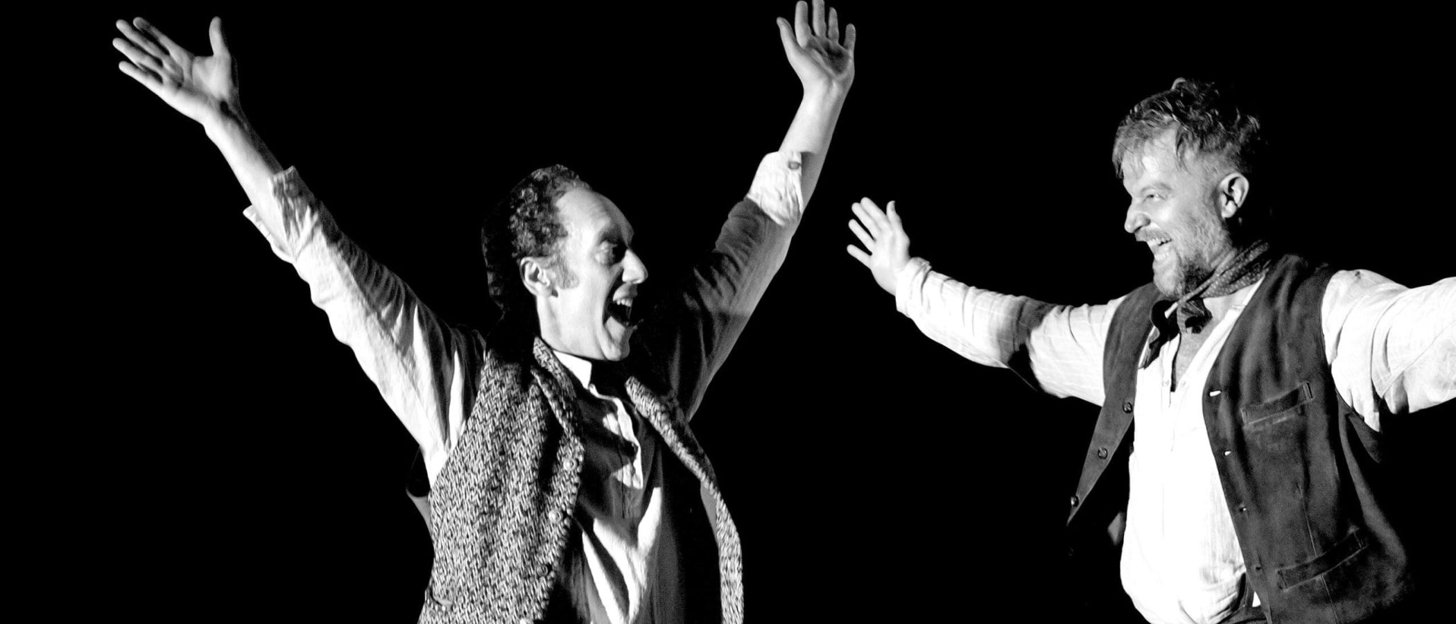 Two men in period costume perform a dramatic scene, arms raised.