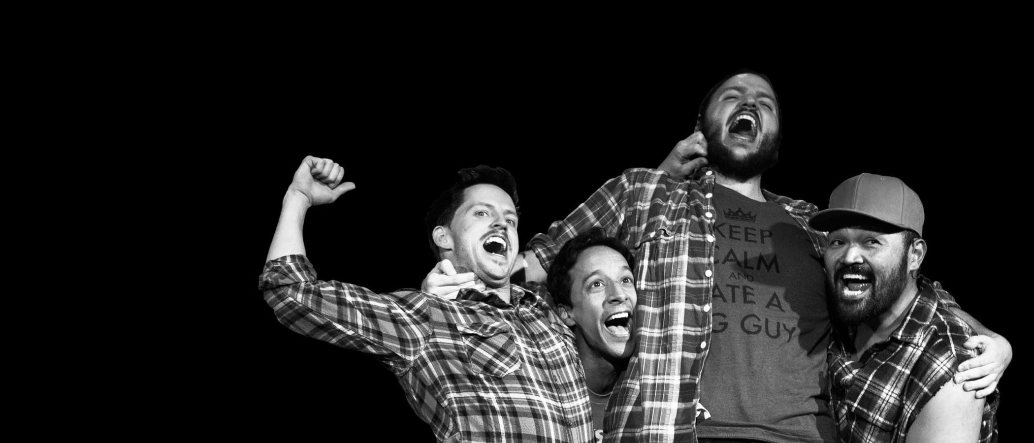 Four joyful men in plaid shirts, one wearing a "Keep Calm and Ate a Big Guy" shirt.