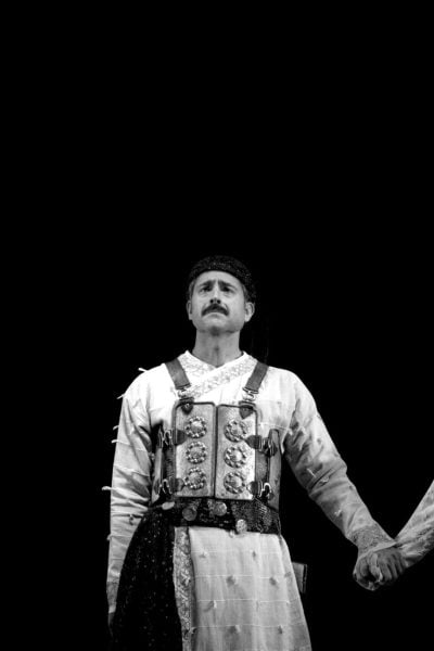 Two guards in traditional attire stand holding hands, possibly at the Taj Mahal.