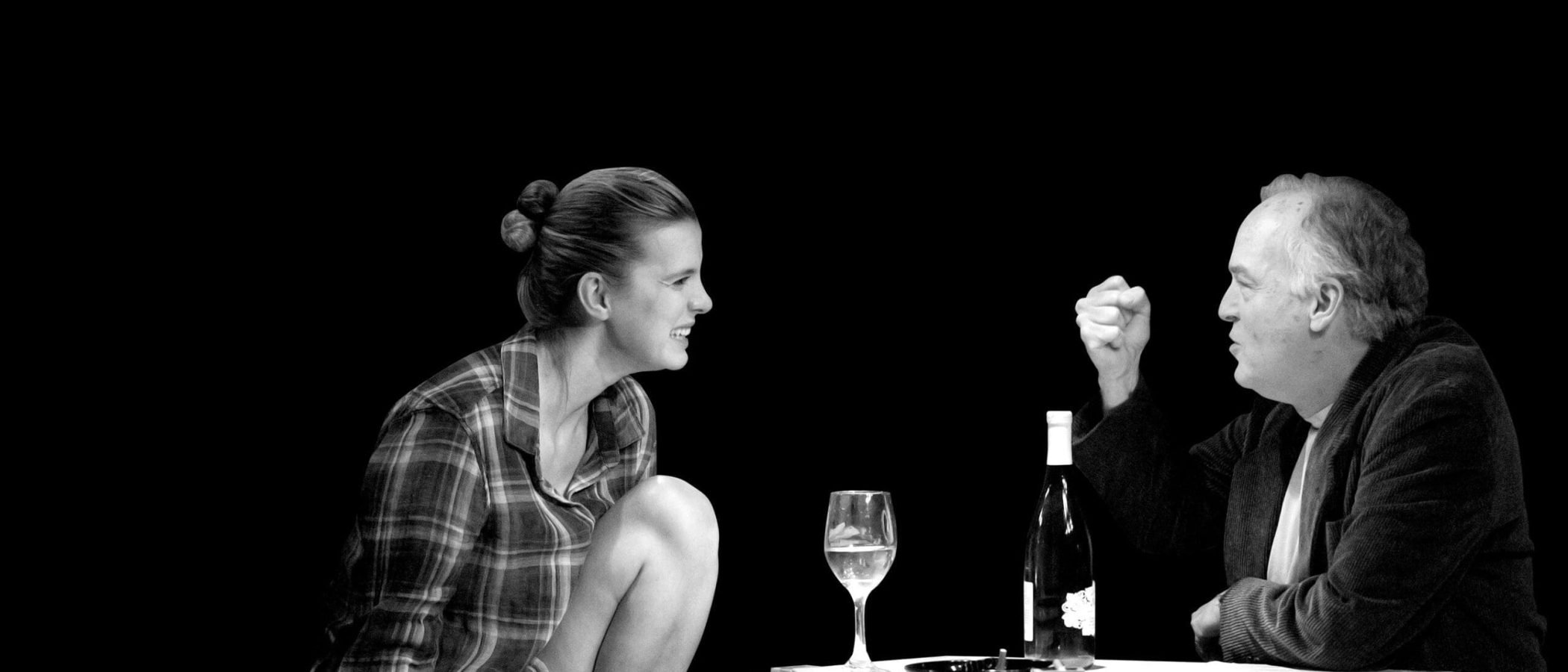 Black and white photo of a man and woman having a tense conversation over wine and cigarettes.