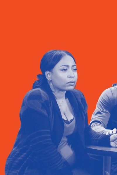 A woman and man sit at a table, holding hands. They appear concerned.