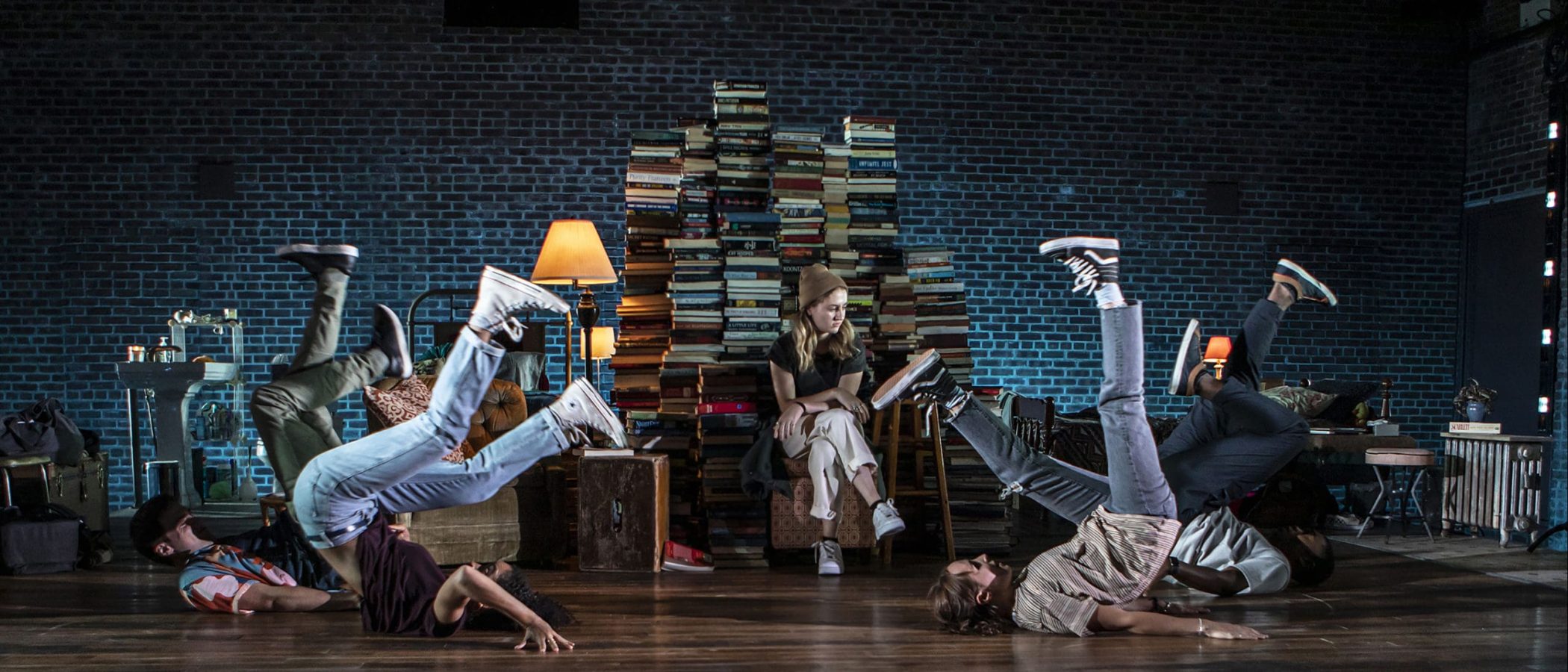 2700x1800_Sunday_Hero-scaled Four dancers breakdancing on a stage, with a woman seated calmly amidst books in the background.