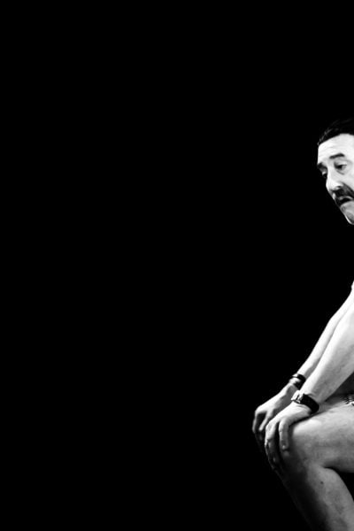 Man sits pensively on a stool against a black background.