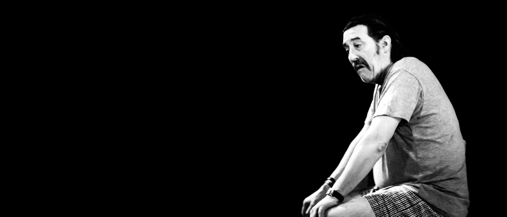 2700x1800_The-Night-Alive_1-scaled Man sits pensively on a stool against a black background.