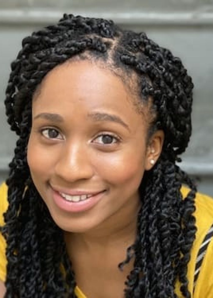 Angel Adedokun smiling, wearing a yellow shirt with black twist braids