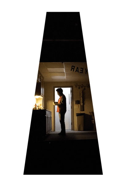 Man standing in dimly lit office breakroom, drinking from a mug.