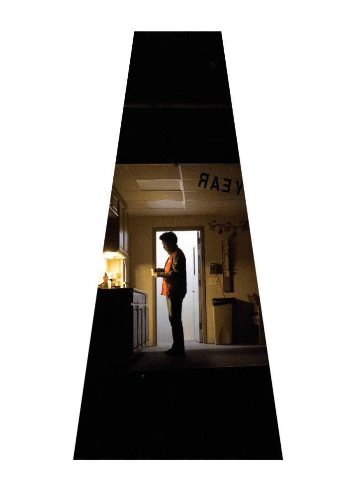 Man standing in dimly lit office breakroom, drinking from a mug.