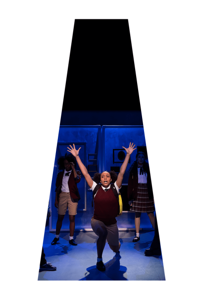 A young woman in a school uniform kneels center stage, arms raised, during a performance of *She Persisted, The Musical*.