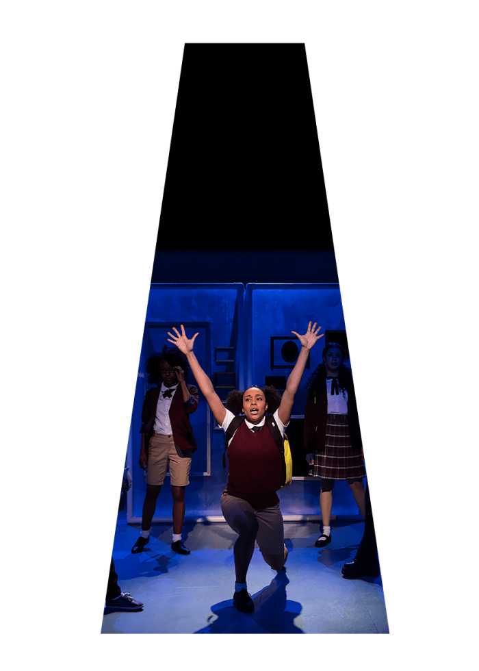 A young woman in a school uniform kneels center stage, arms raised, during a performance of *She Persisted, The Musical*.
