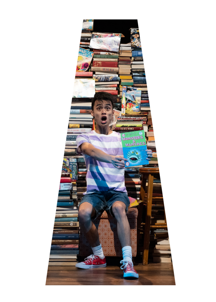 Young actor, surprised expression, holding "Monsters & Maniacs" book, surrounded by books.