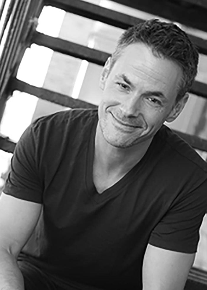 Adam Guettel smiling, seated, wearing a dark v-neck shirt