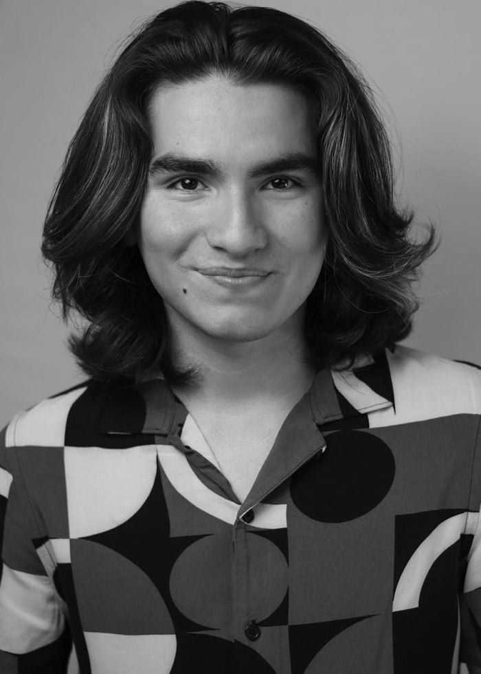 Young man with long hair smiling, wearing a patterned shirt.