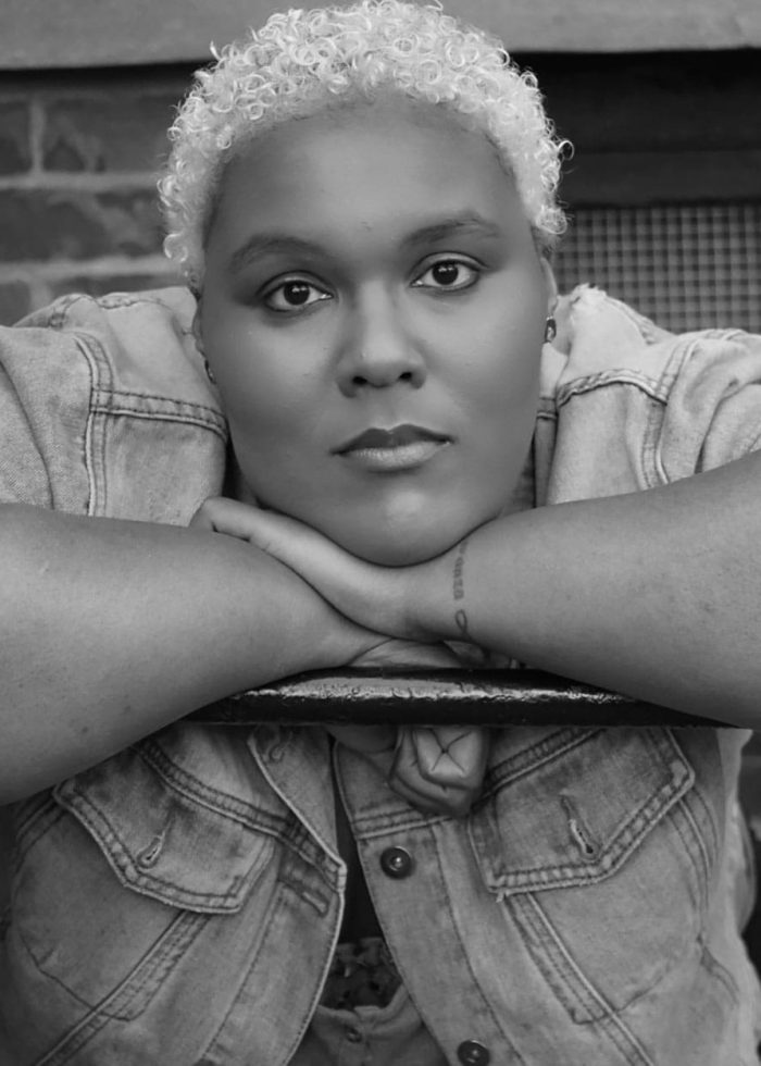 Amara Brady, a woman with short, curly blonde hair, leans on her forearms, gazing at the camera.