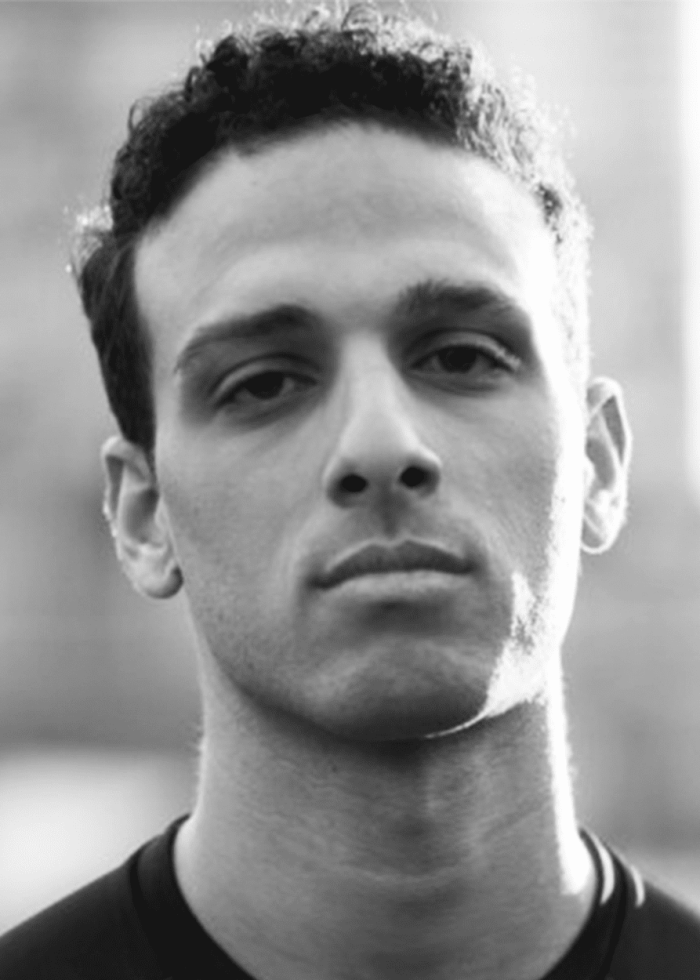 Ari'el Stachel, a man with dark curly hair, looking directly at the camera in black and white.