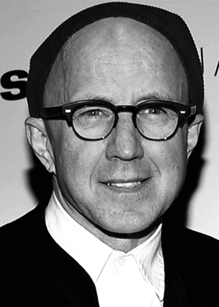 Arliss Howard in a black and white portrait, wearing glasses and a beanie, smiling.