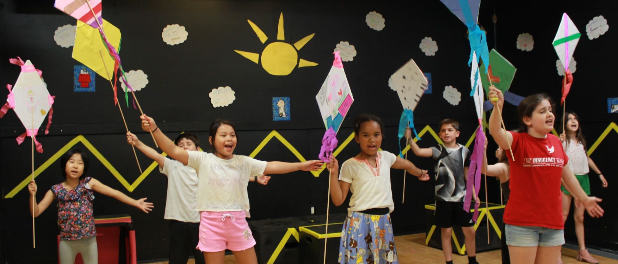 Children on stage with kites in a theatrical performance, bright backdrop with sun.