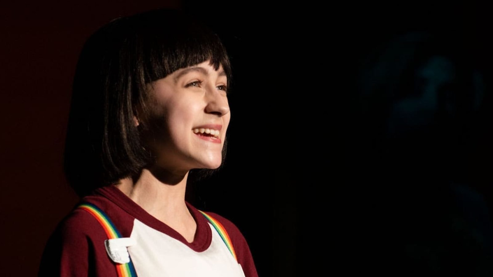 Smiling young woman with short dark hair, wearing a maroon shirt and rainbow suspenders.