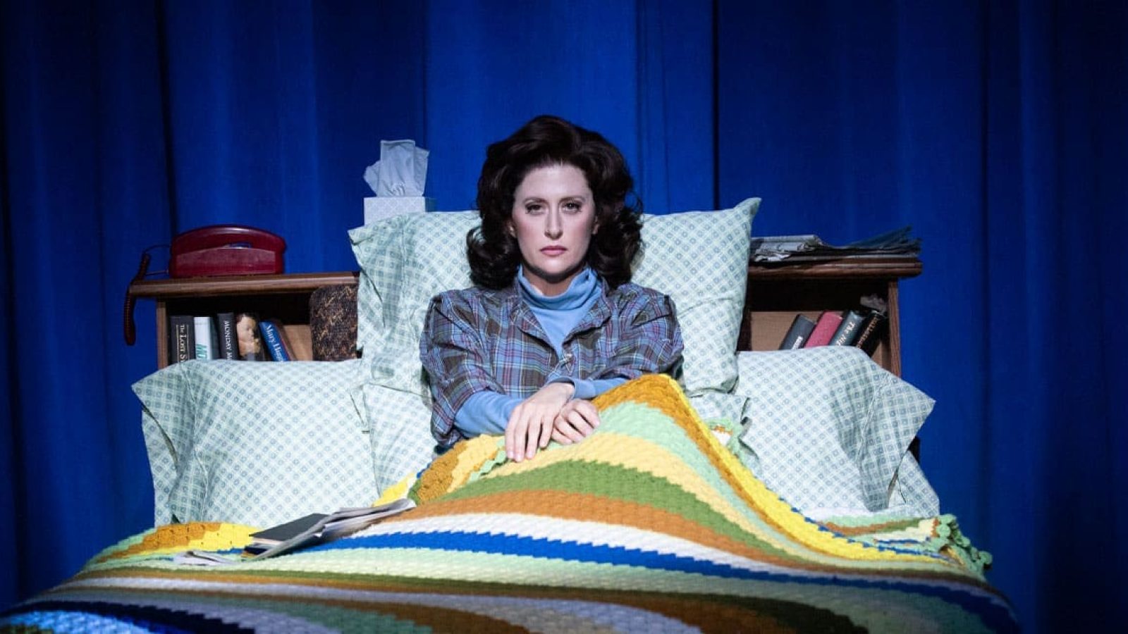 Woman sits in bed, looking pensive, under a colorful blanket.