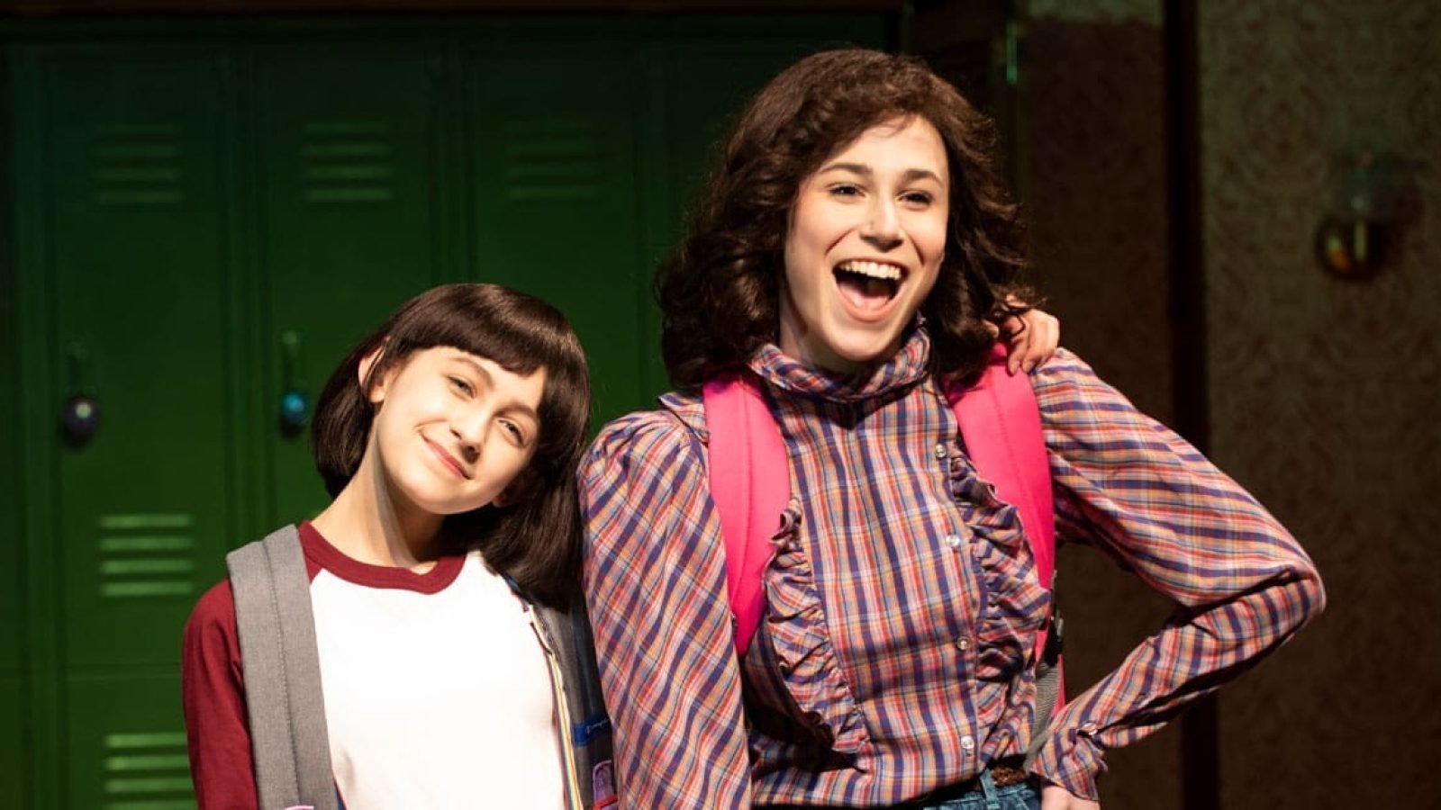 Two young women, one with a bob haircut, stand smiling together in a school hallway. They wear backpacks.