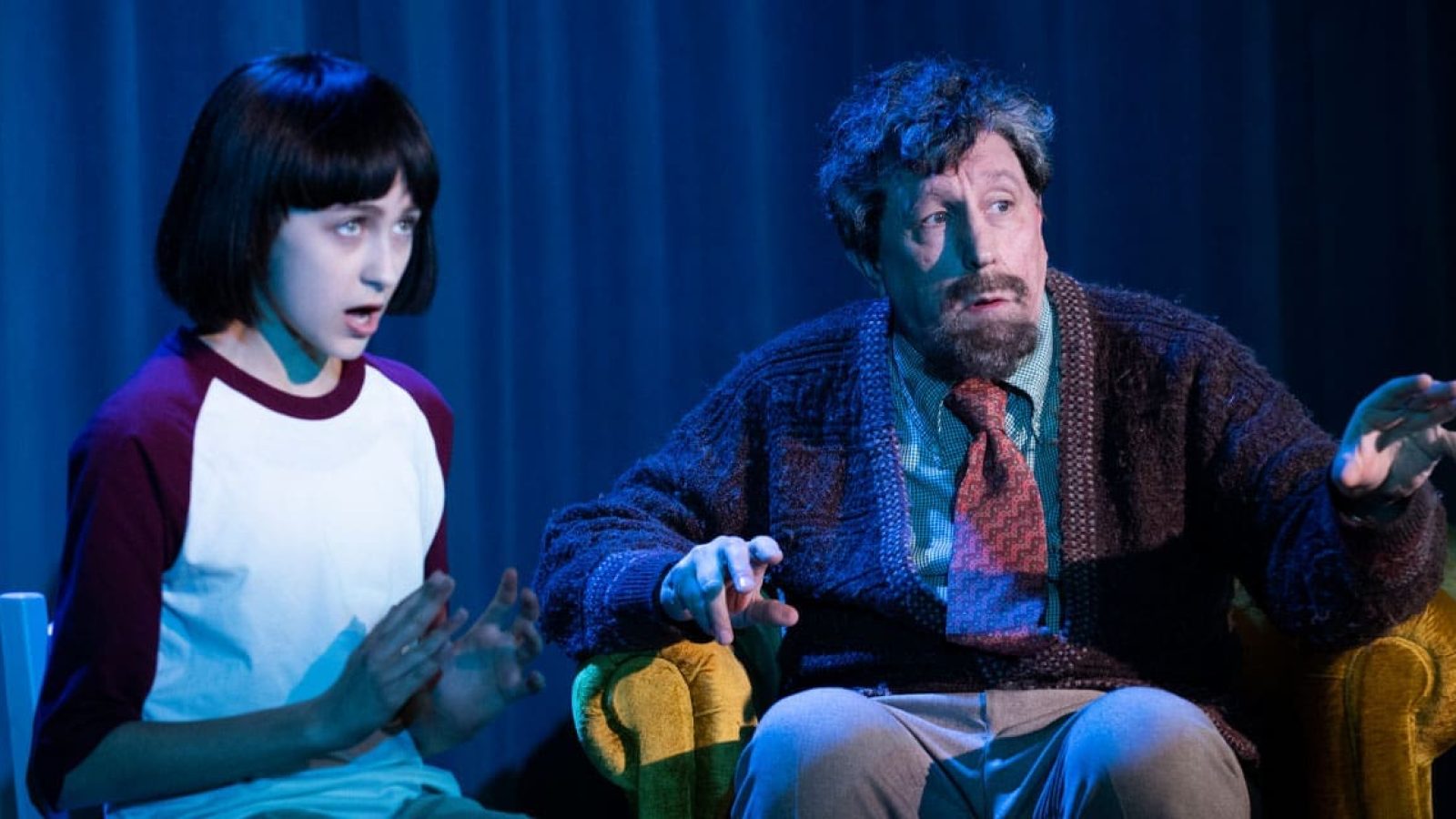 A young girl and older man sit on a couch, engaged in conversation. The girl appears concerned.