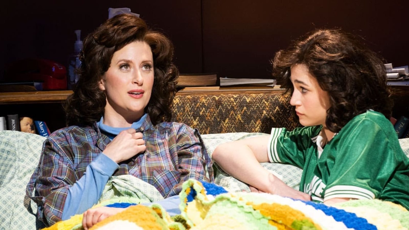 Two women, one in a plaid shirt and the other in a green jacket, share a quiet moment in bed. This is a scene from the play, *The Bedwetter*.
