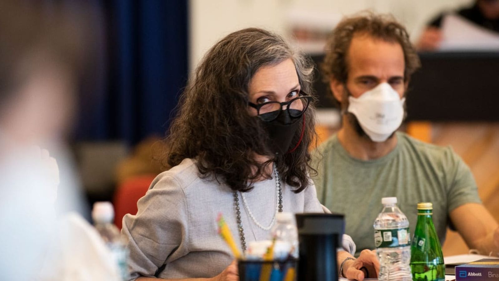 Two masked individuals, a woman and a man, sit at a table during a meeting.