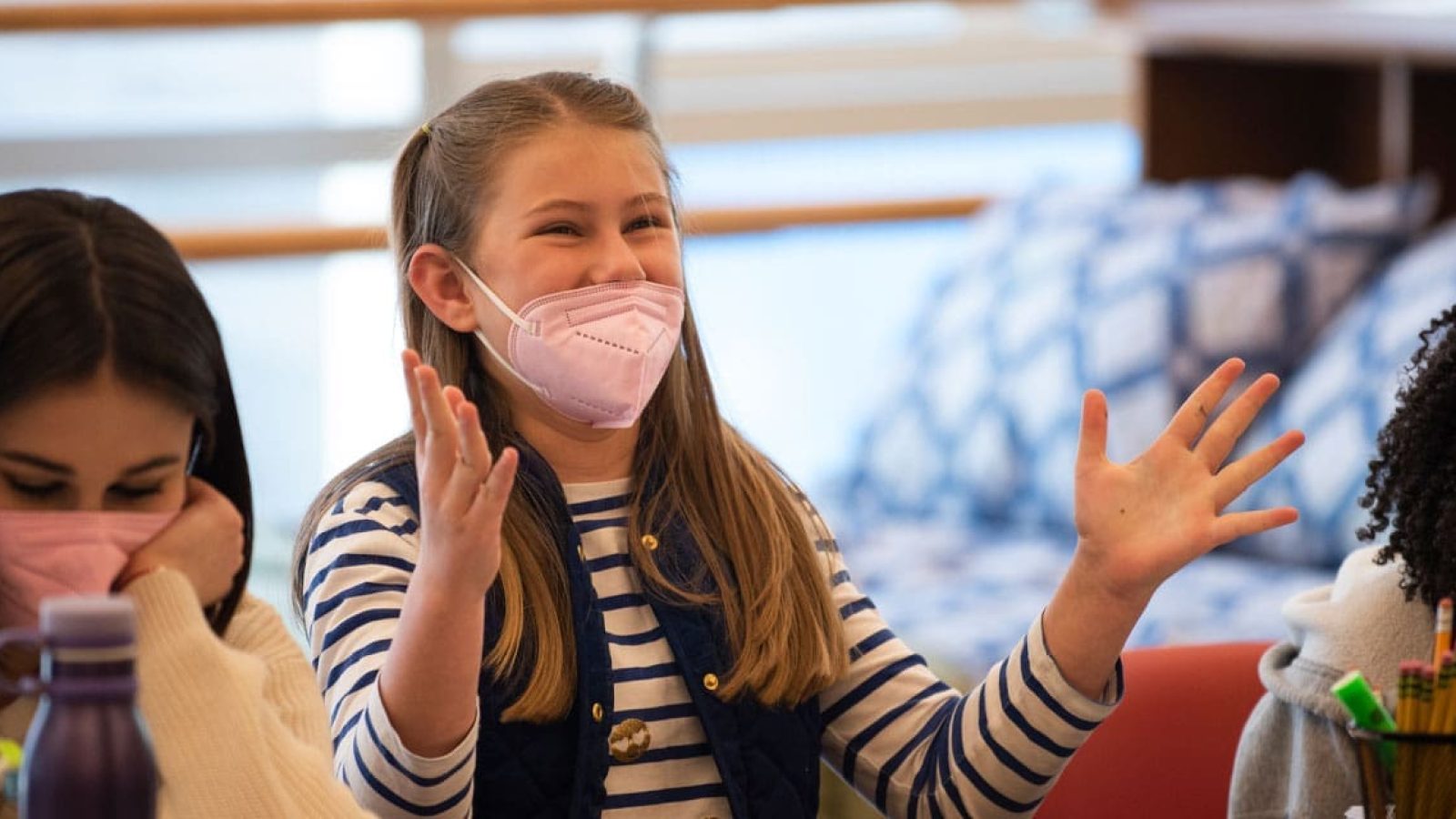 Three girls in a classroom, one raises her hand enthusiastically.