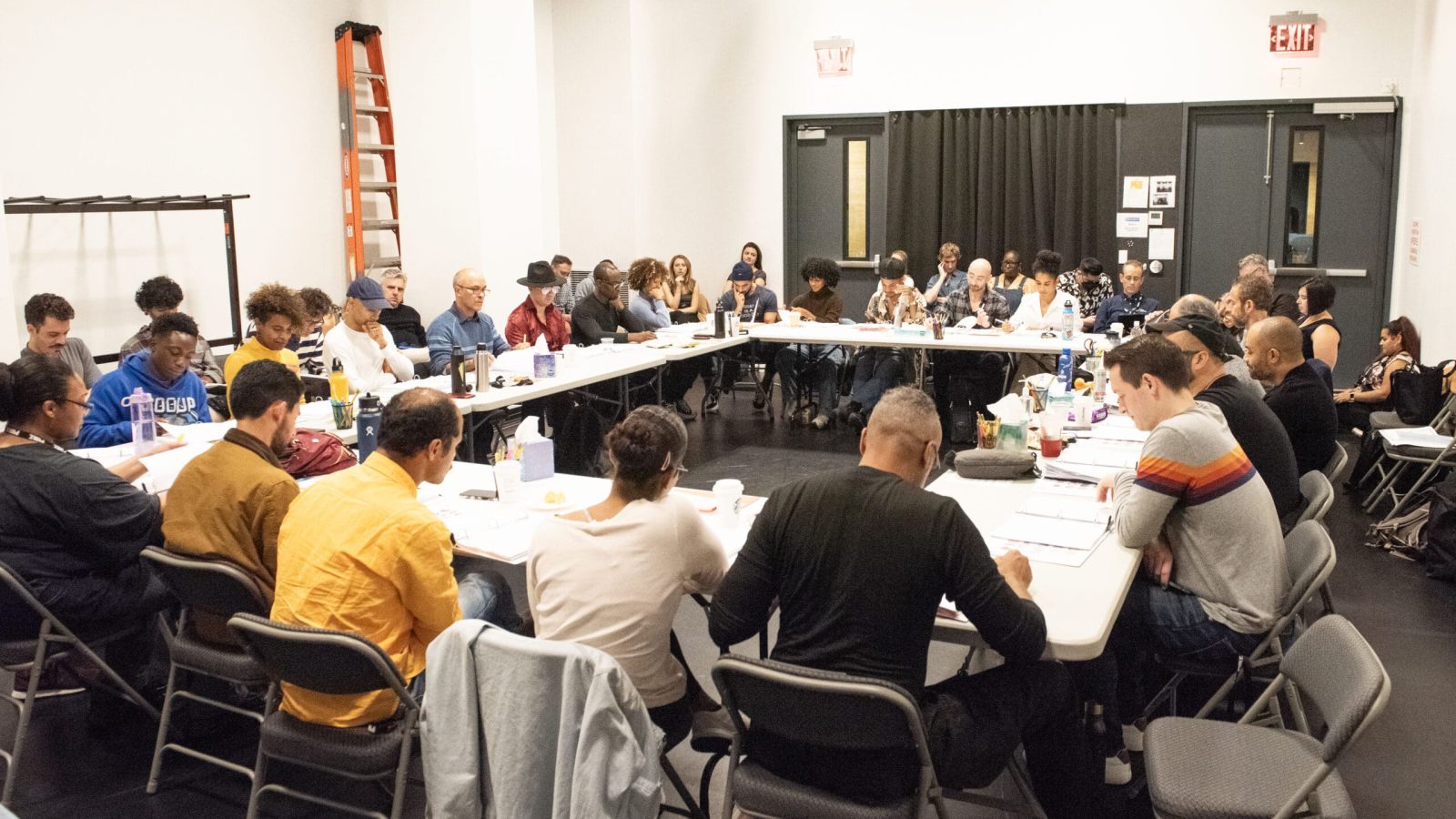 A large group of people sits around tables in a room, engaged in a meeting or discussion.