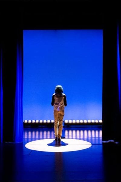 A lone figure stands center stage, back to the camera, illuminated by a spotlight against a blue backdrop.