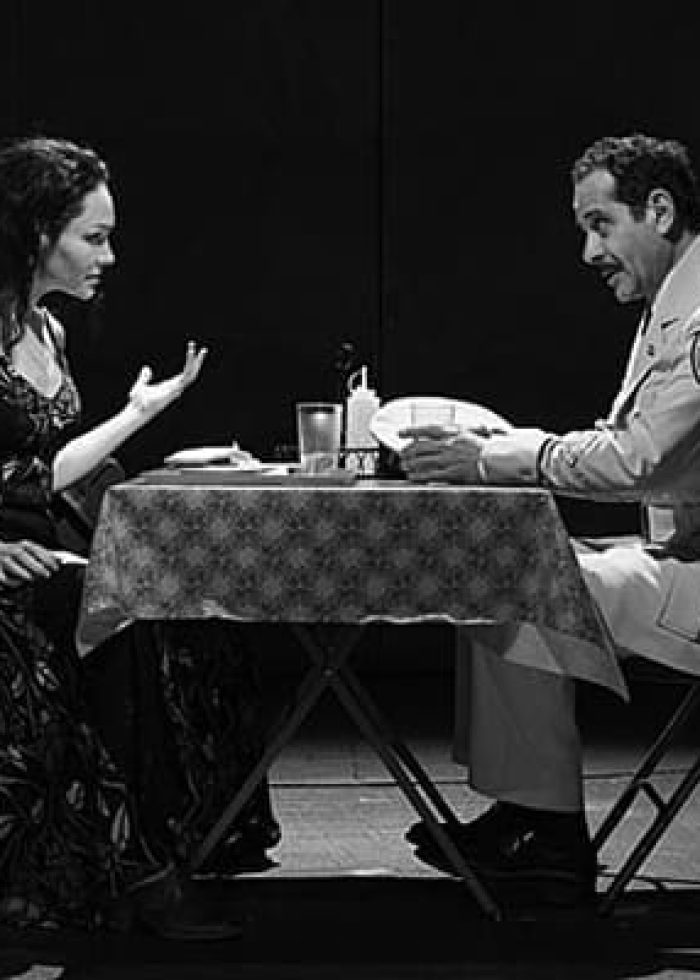 Actors in a tense scene. A woman and a man in uniform sit at a table, possibly discussing a play.