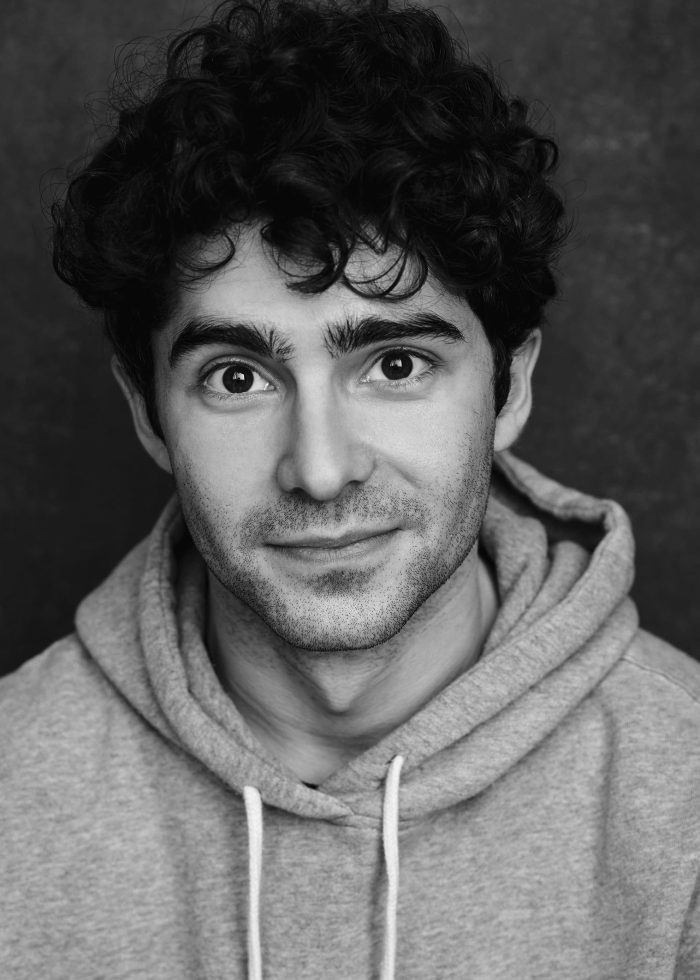 Young man with curly hair smiles, wearing a grey hoodie