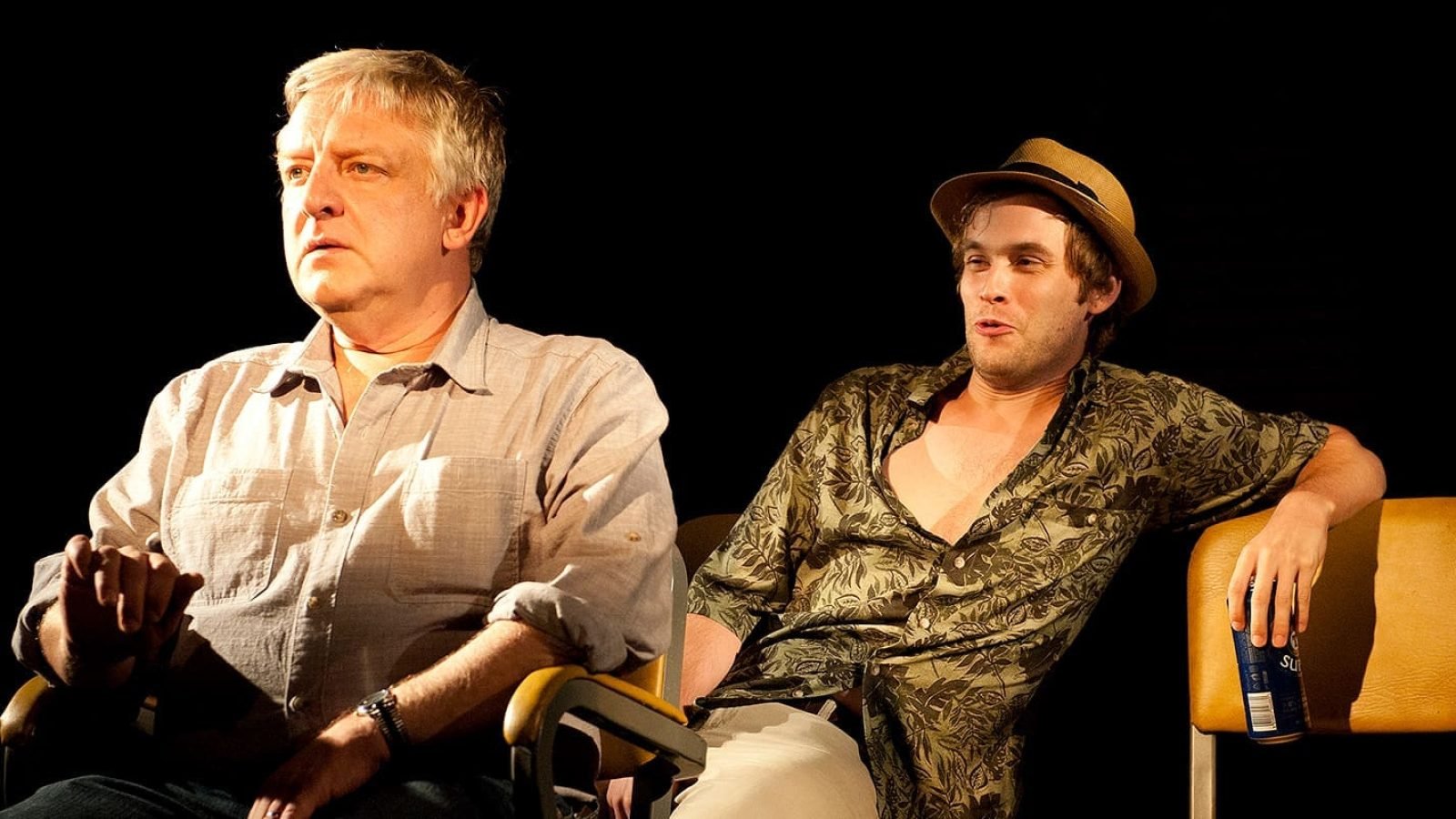 Two men sitting in chairs, one wearing a floral shirt and hat.