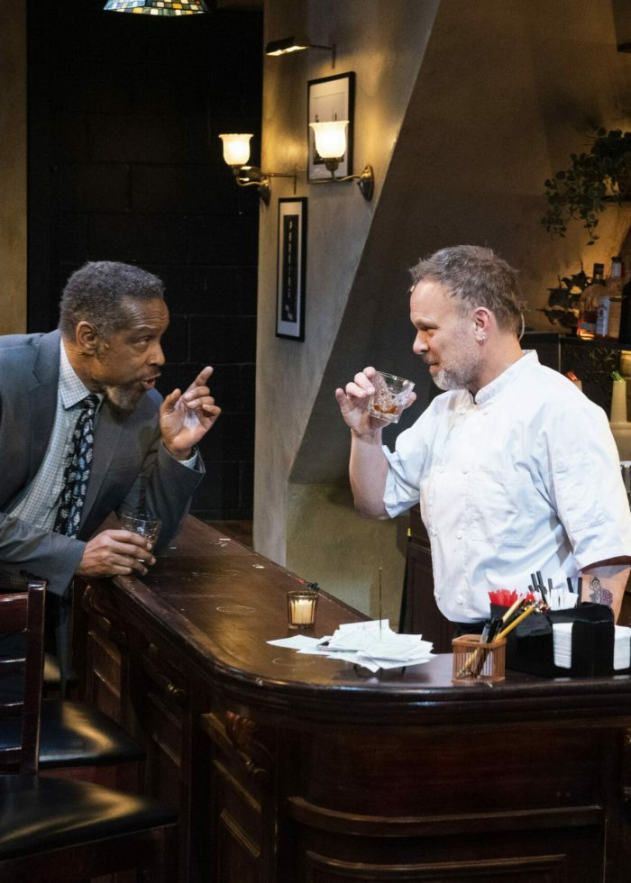 Two men in a bar, one in a suit gesturing, the other a chef holding a drink. Scene from 'Cornelia Street'.