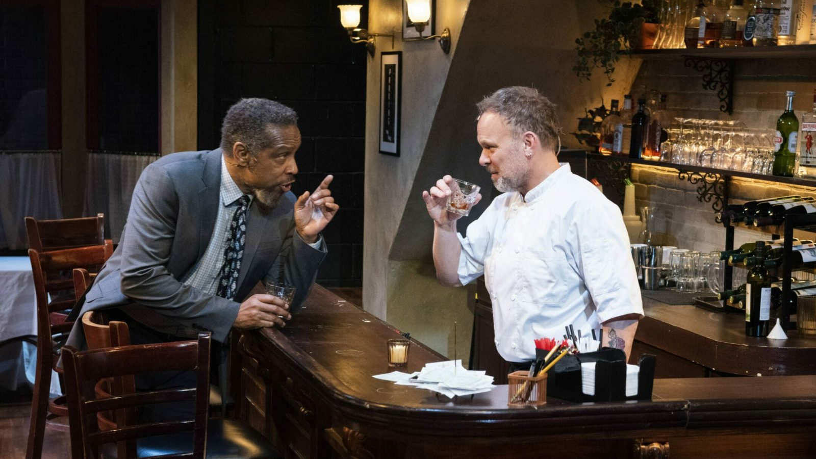 Two men in a bar, one in a suit gesturing, the other a chef holding a drink. Scene from 'Cornelia Street'.