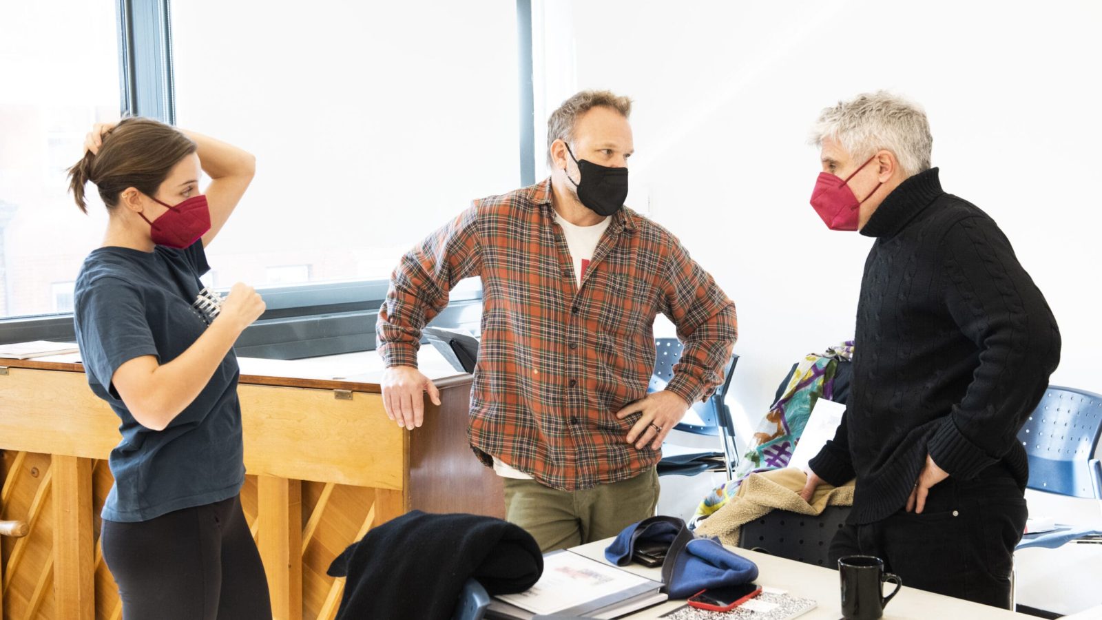 Three masked individuals converse in a room; one woman, two men.
