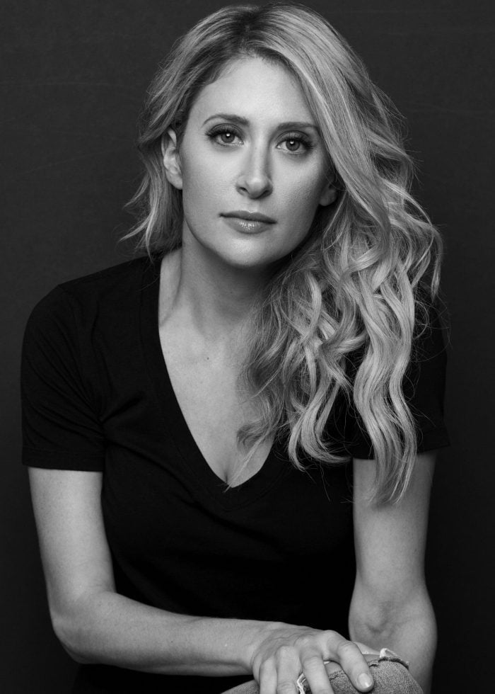 Caissie Levy in a black v-neck shirt, looking at the camera with wavy blonde hair.