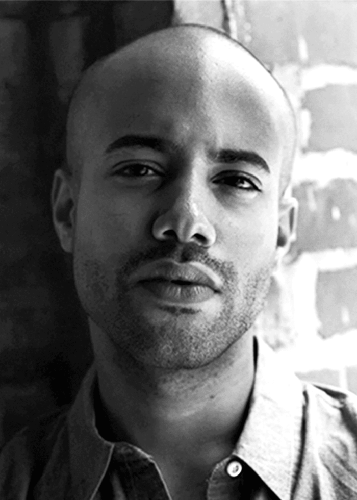 Sean Carvajal, a man with a shaved head, gazes directly at the camera, leaning against a brick wall.