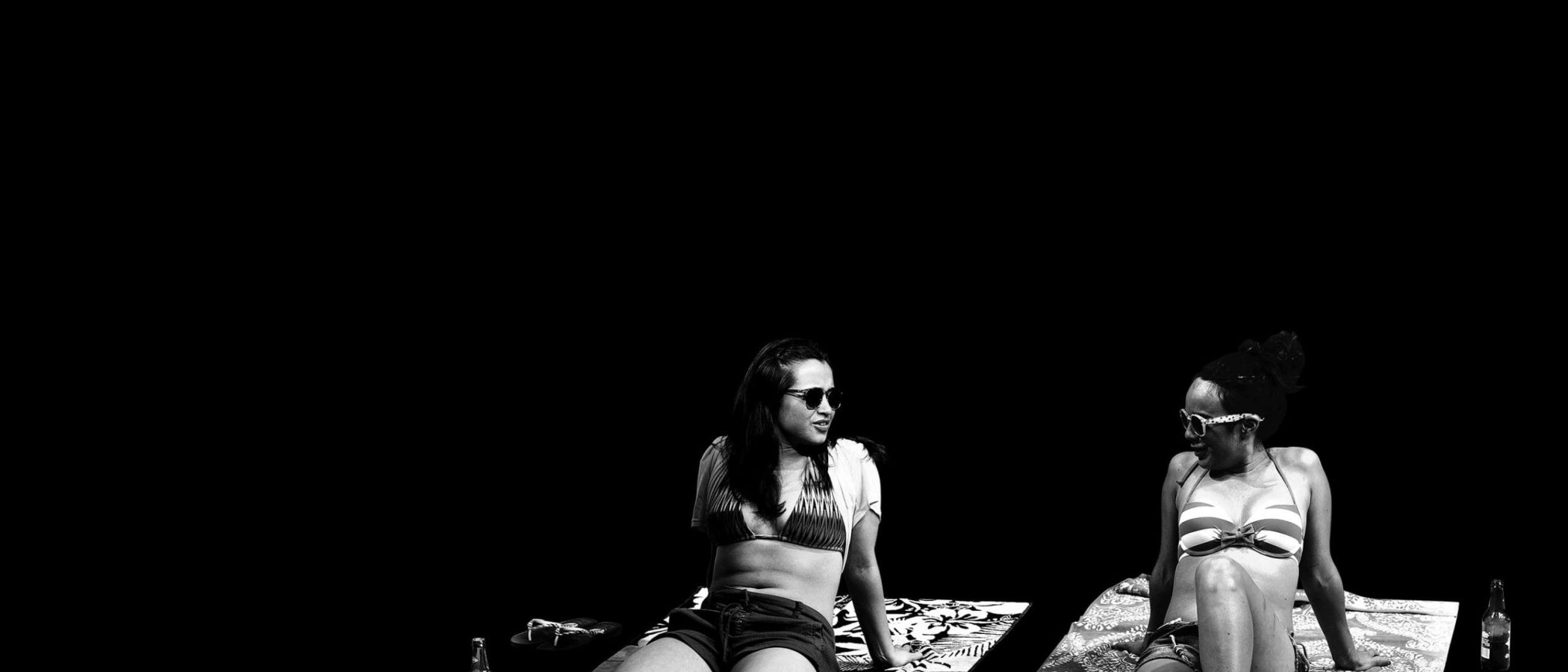 Two women relax on beach towels, wearing sunglasses and bikinis.